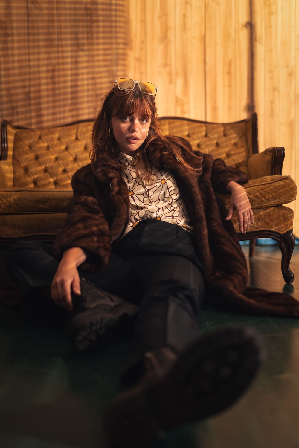 Woman in brown fur coat and sunglasses sitting on floor against vintage velvet sofa.