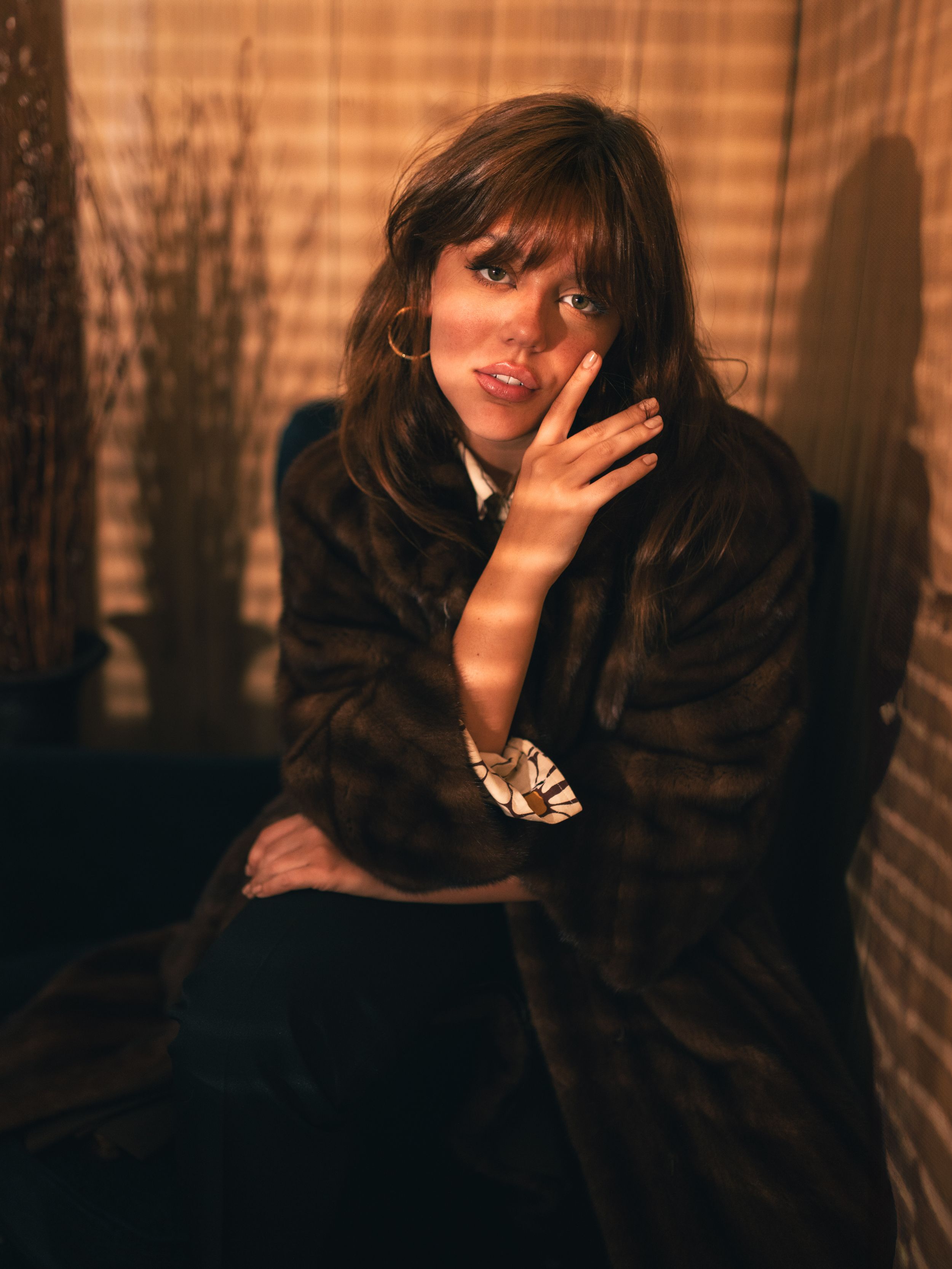 Brunette woman in dark fur coat posing thoughtfully in warm ambient lighting.