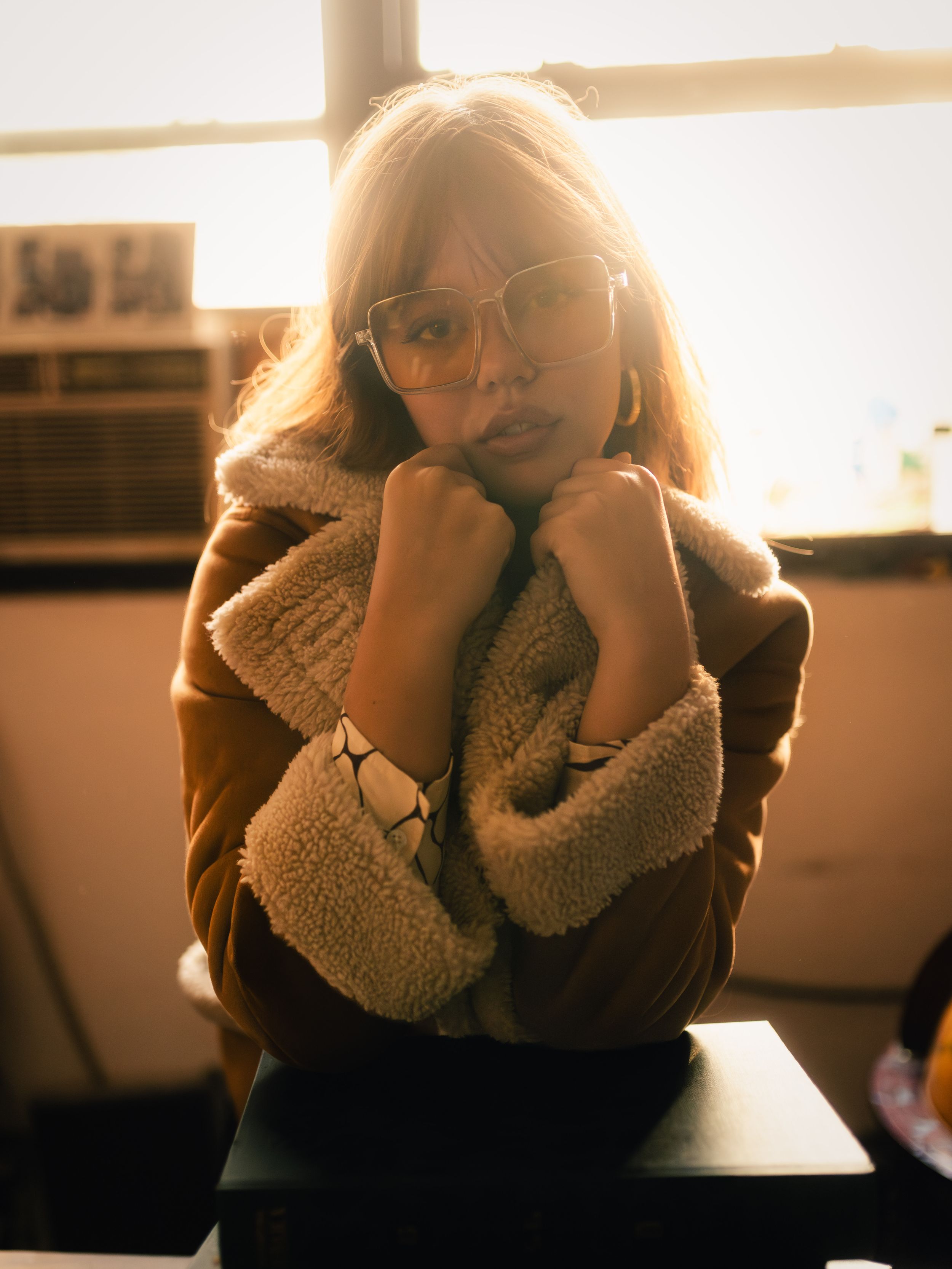 Young woman in brown shearling jacket and glasses poses indoors in warm golden backlight.