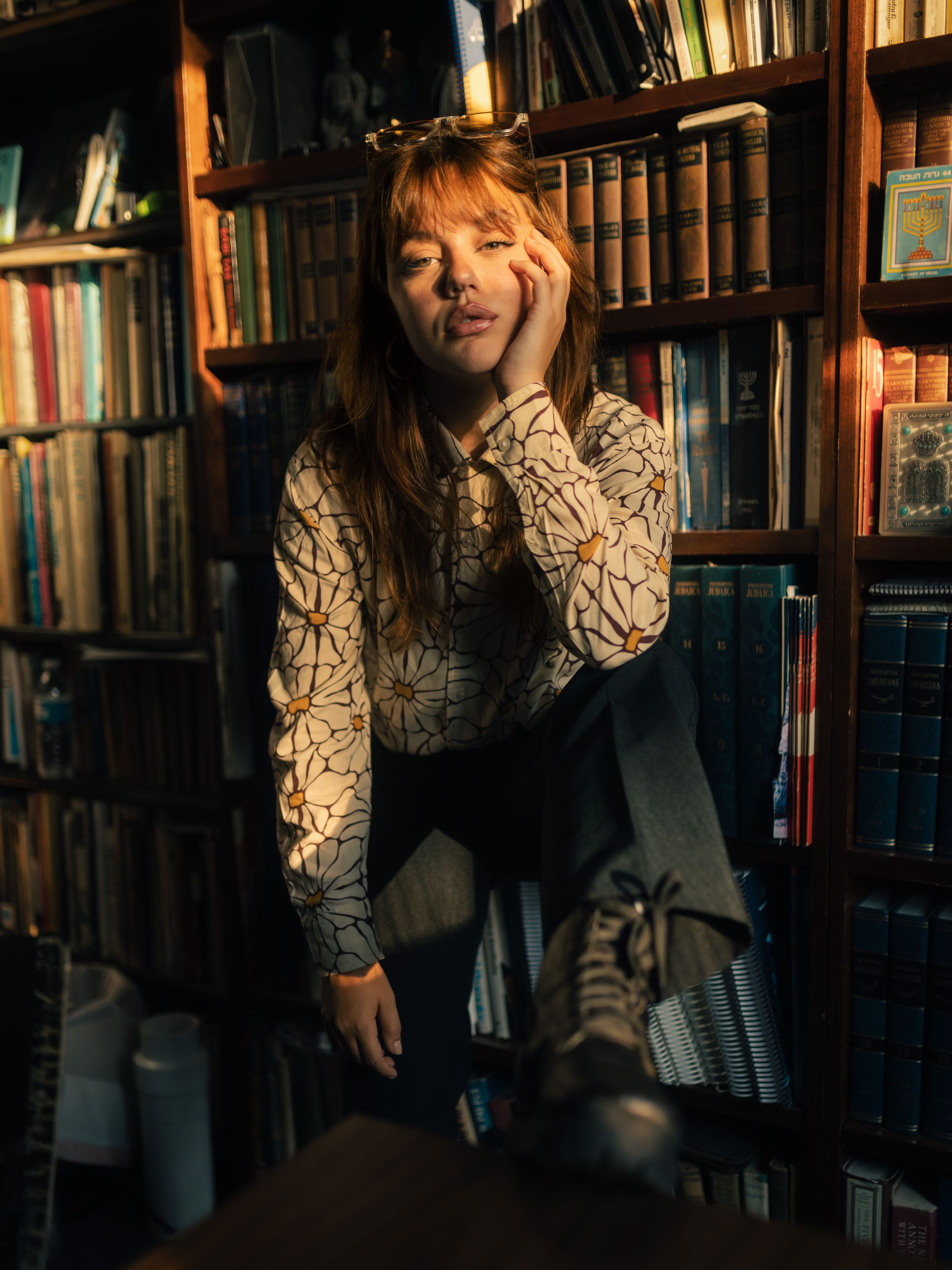Young woman in floral blouse poses thoughtfully among warm-lit bookshelves in a cozy library setting.