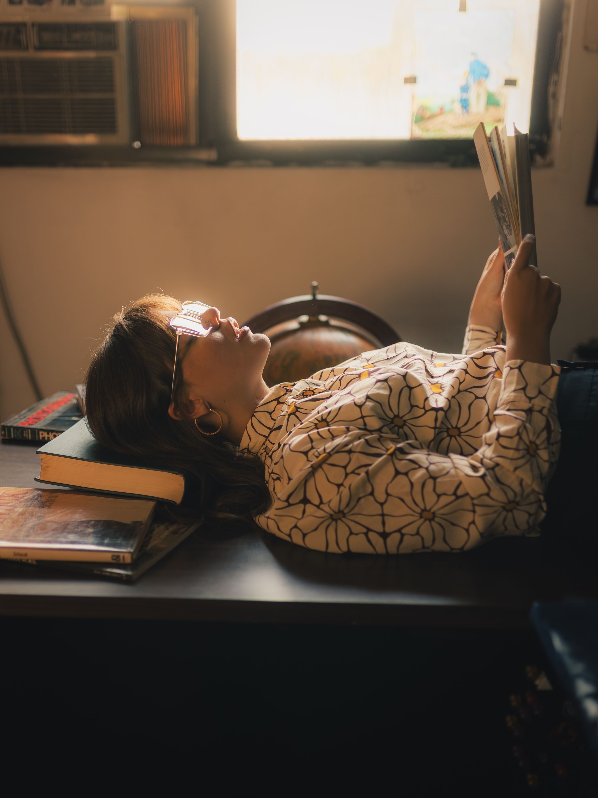 A young woman lying on a desk reading a book, resting her head on stacked books in warm sunlight.