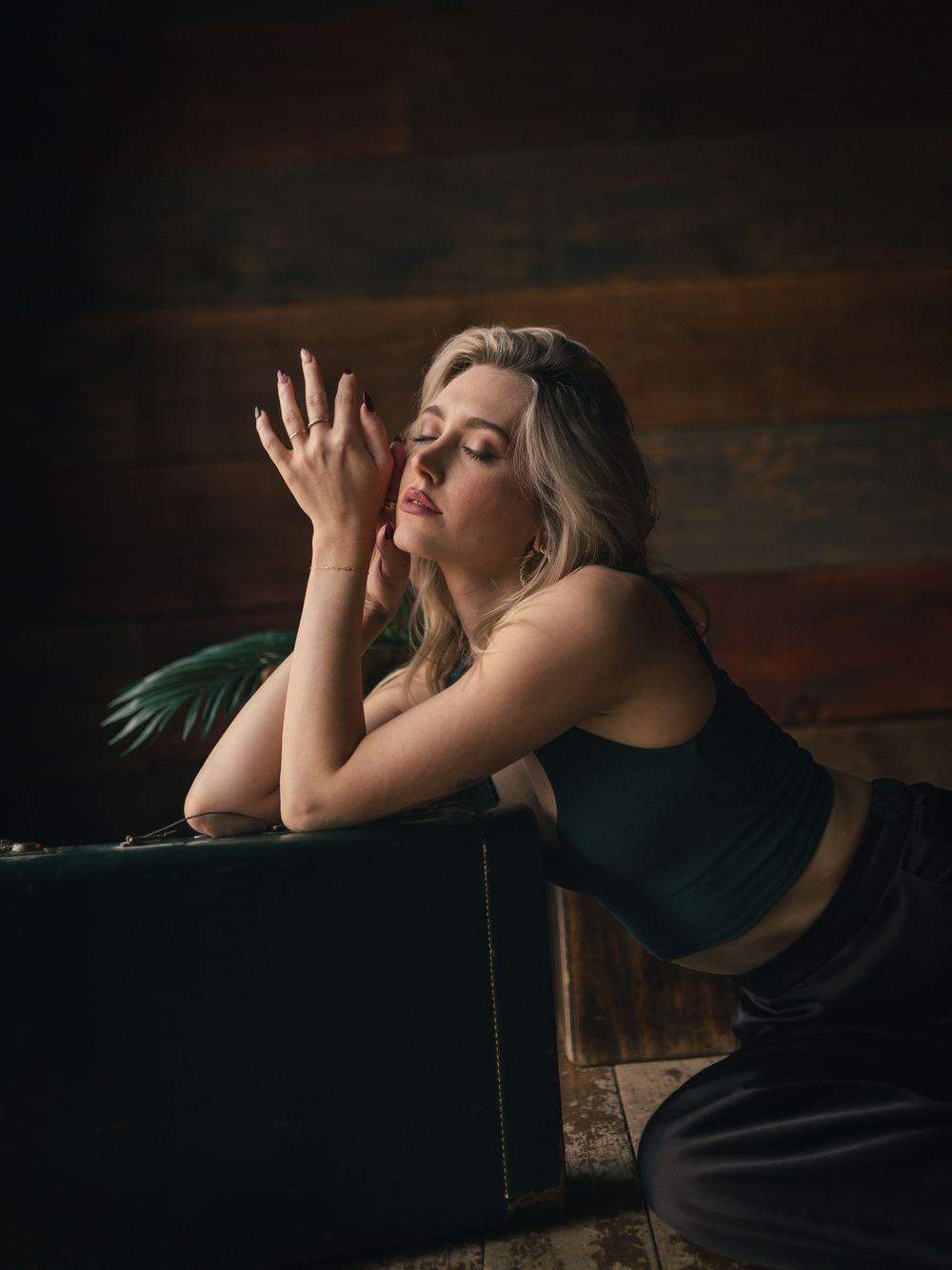 Blonde woman in green crop top poses dramatically leaning on vintage suitcase against rustic wood wall in moody lighting.