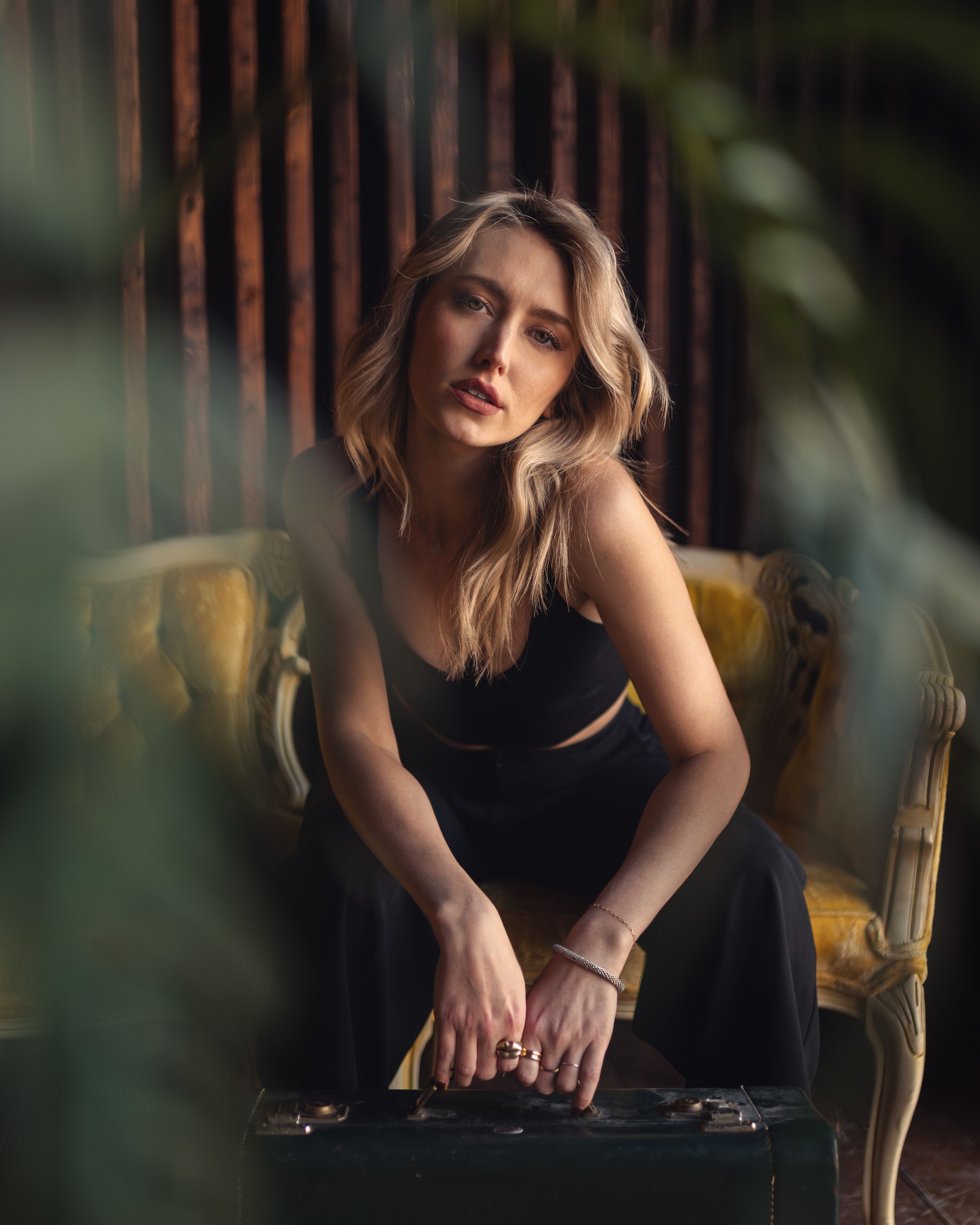 A blonde woman in a black dress sits pensively on a vintage yellow chair in a moody, plant-filled setting.