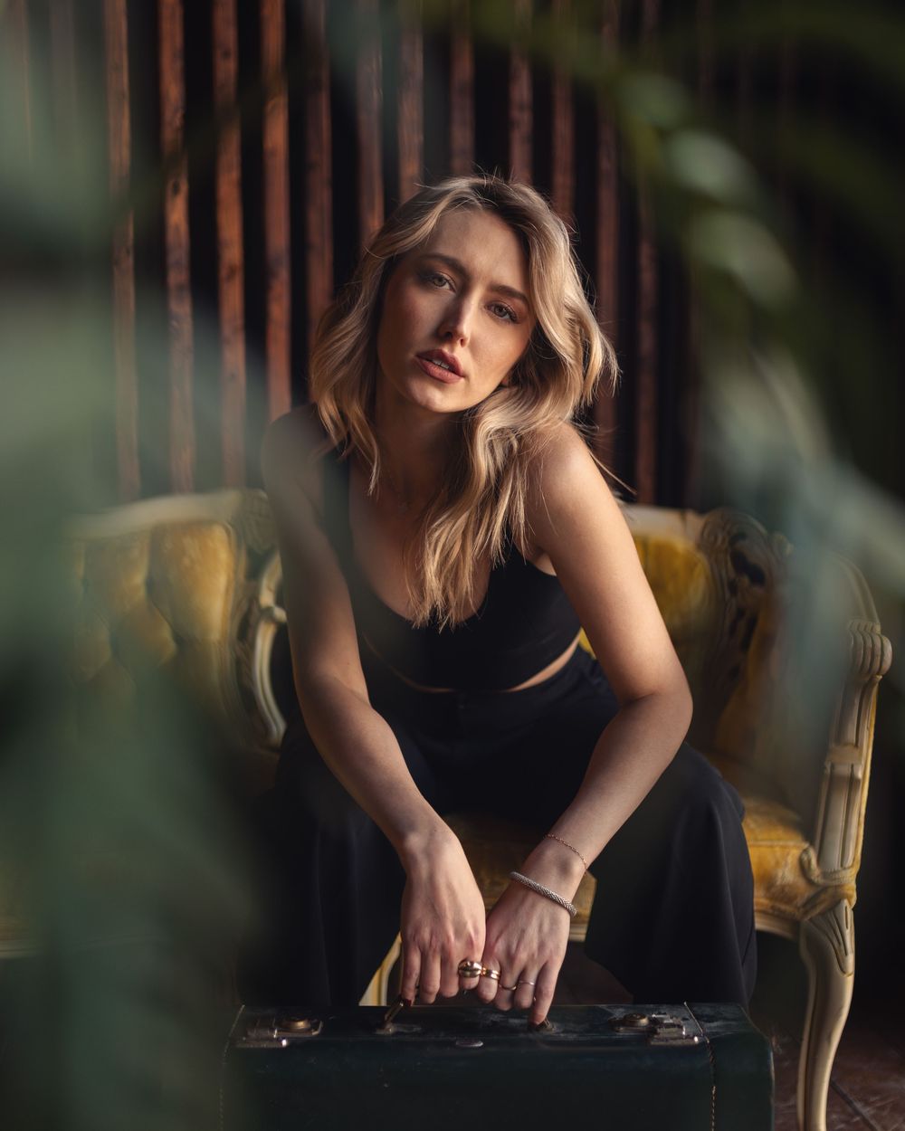 A blonde woman in a black dress sits pensively on a vintage yellow chair in a moody, plant-filled setting.