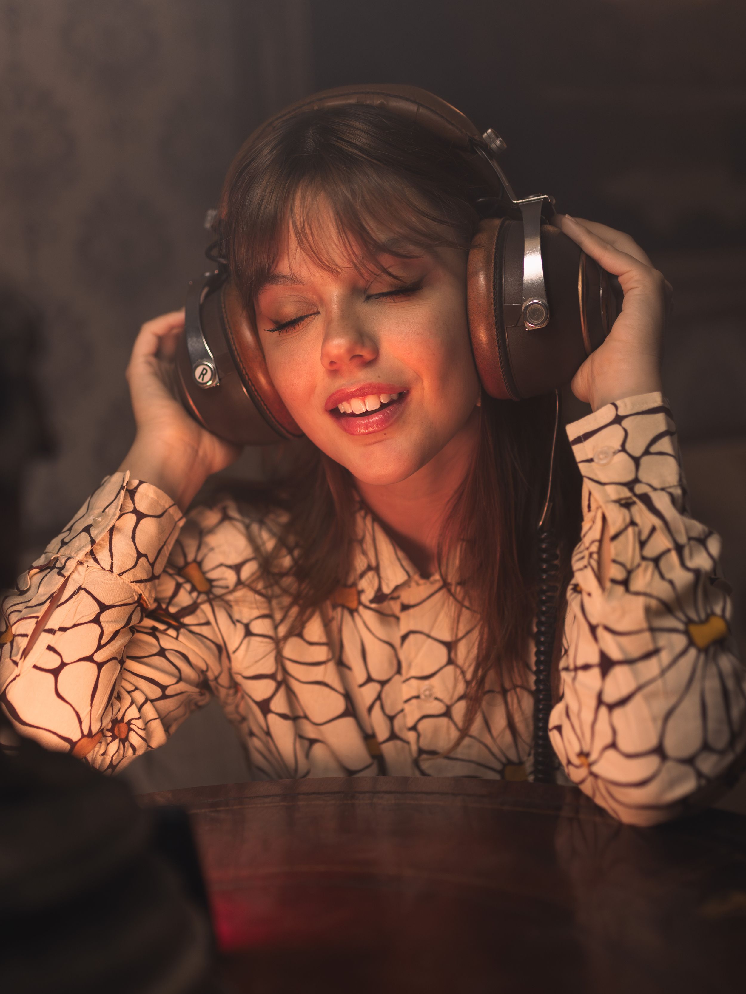 Young woman enjoying music with large headphones, eyes closed, singing along in warm ambient lighting.