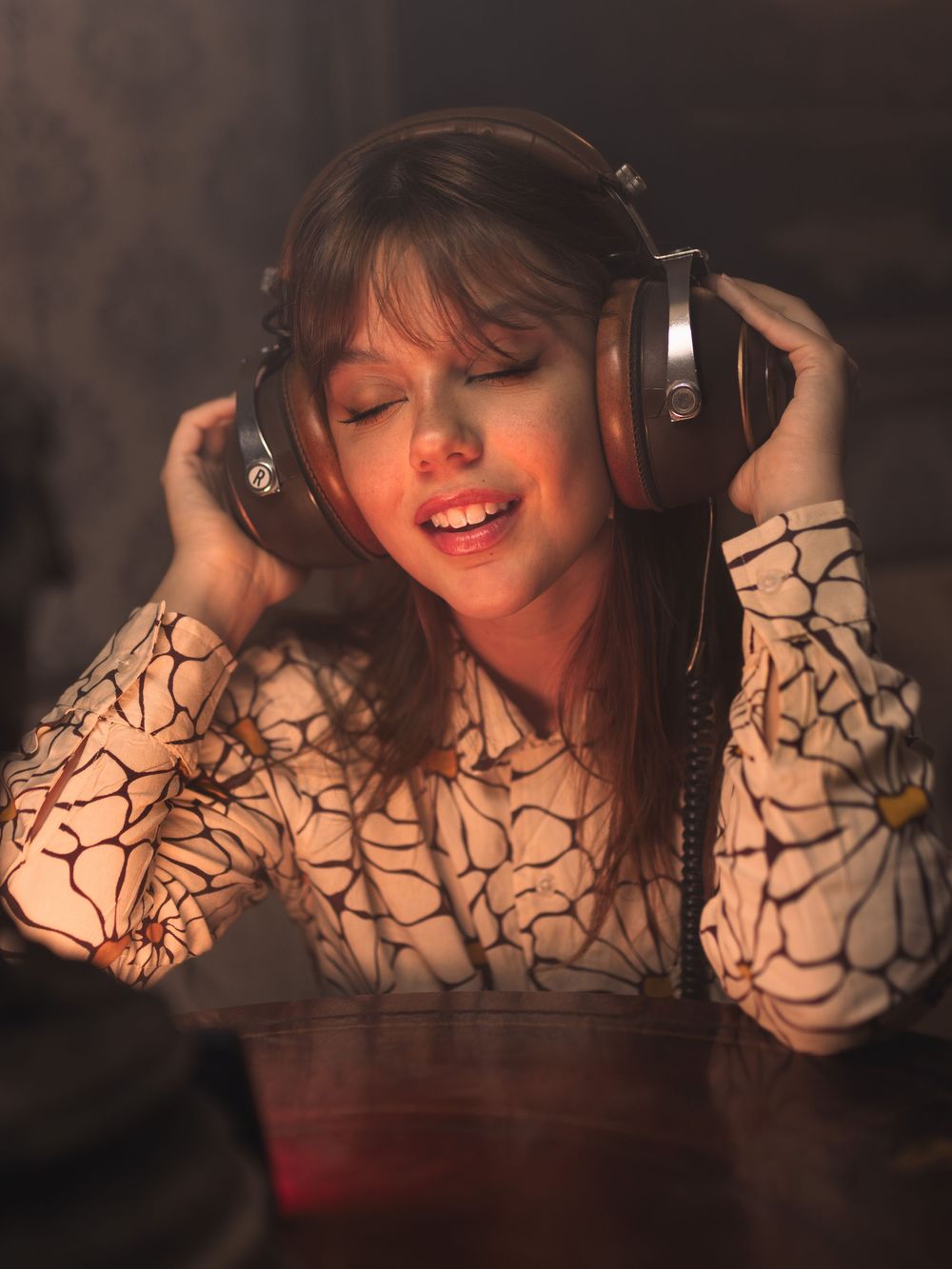 Young woman enjoying music with large headphones, eyes closed, singing along in warm ambient lighting.
