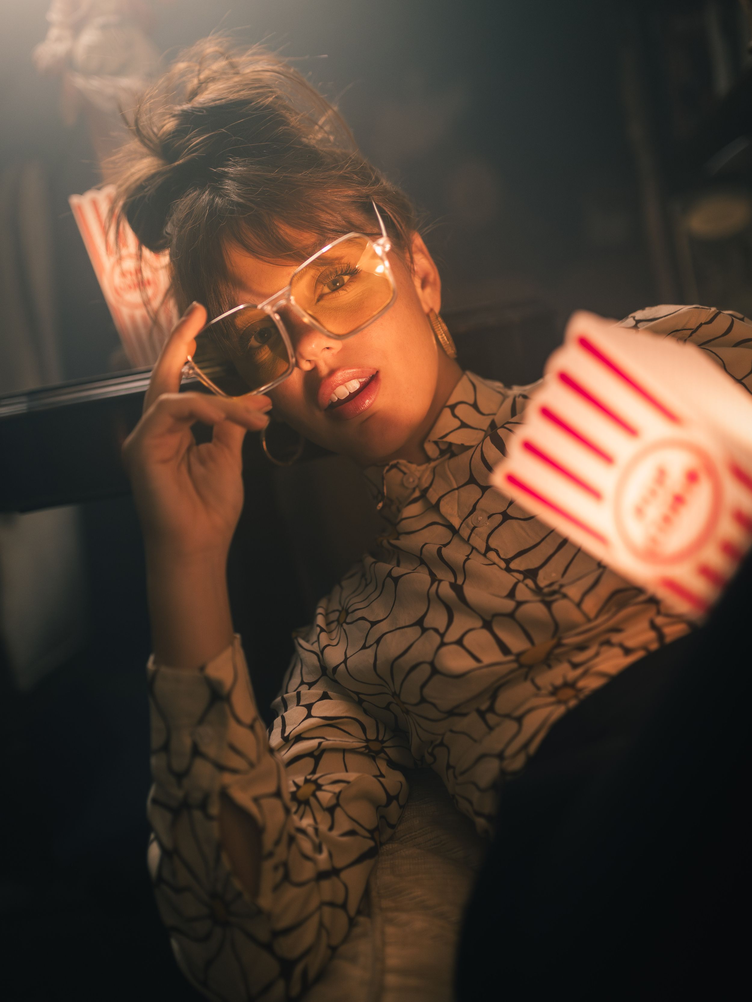 Stylish woman with sunglasses holding popcorn bucket in moody cinema lighting, wearing patterned blouse.