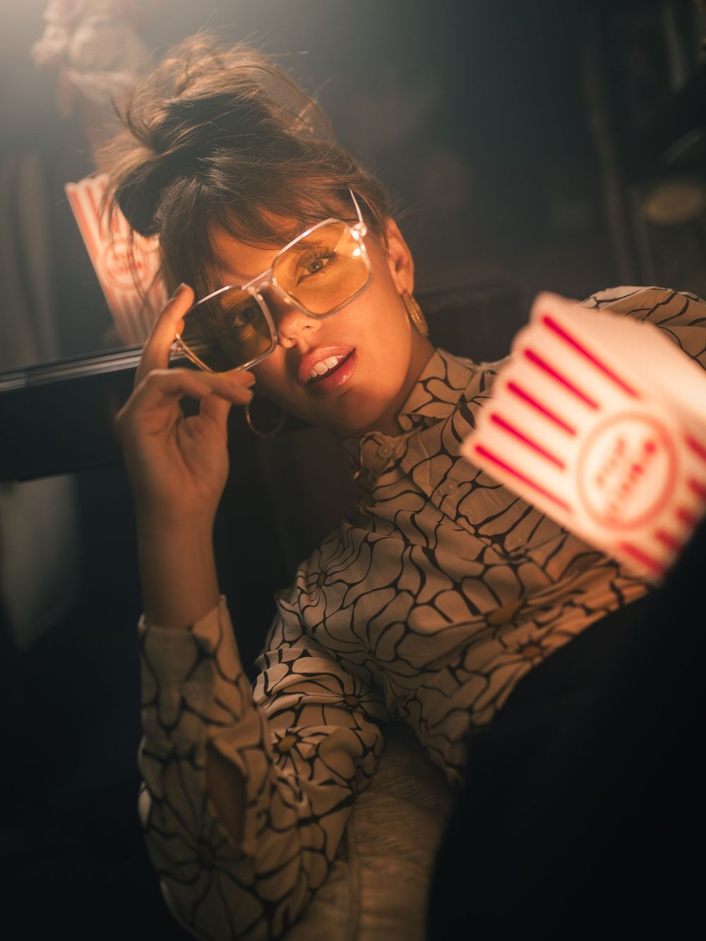 Stylish woman with sunglasses holding popcorn bucket in moody cinema lighting, wearing patterned blouse.