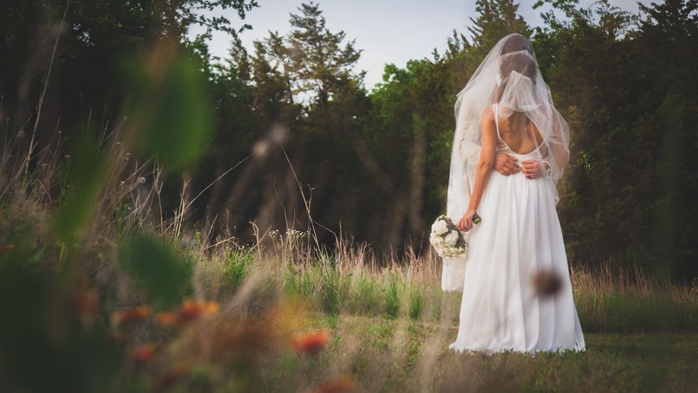Bride in white gown and veil stands in sunlit meadow holding bouquet, surrounded by tall grass and trees.