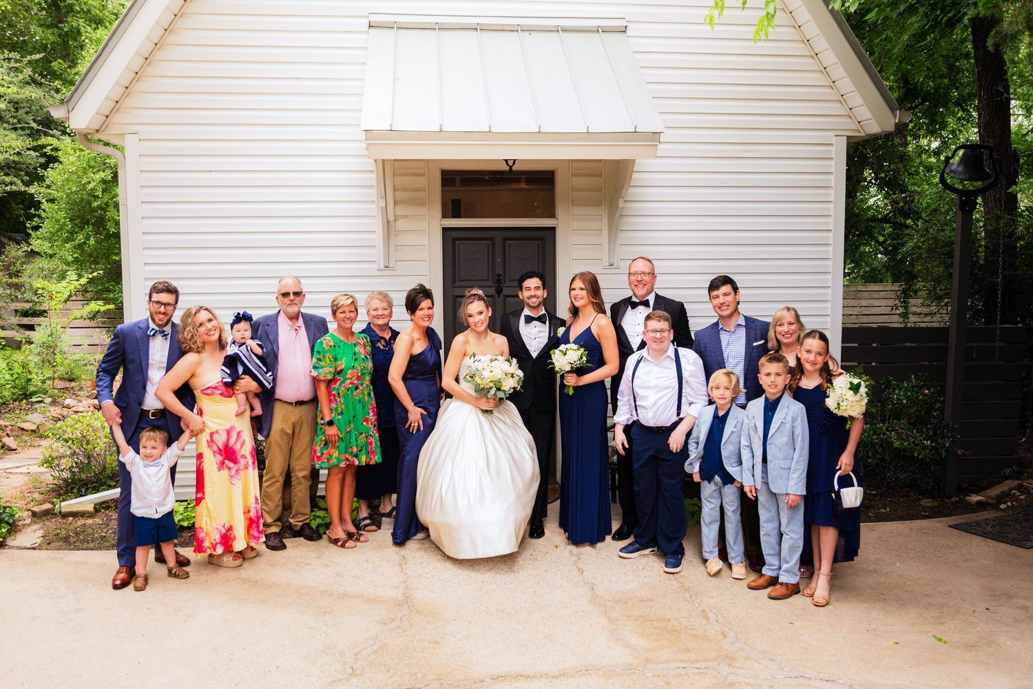 Wedding party poses in front of a white chapel, bride in white gown surrounded by family in navy and colorful attire.