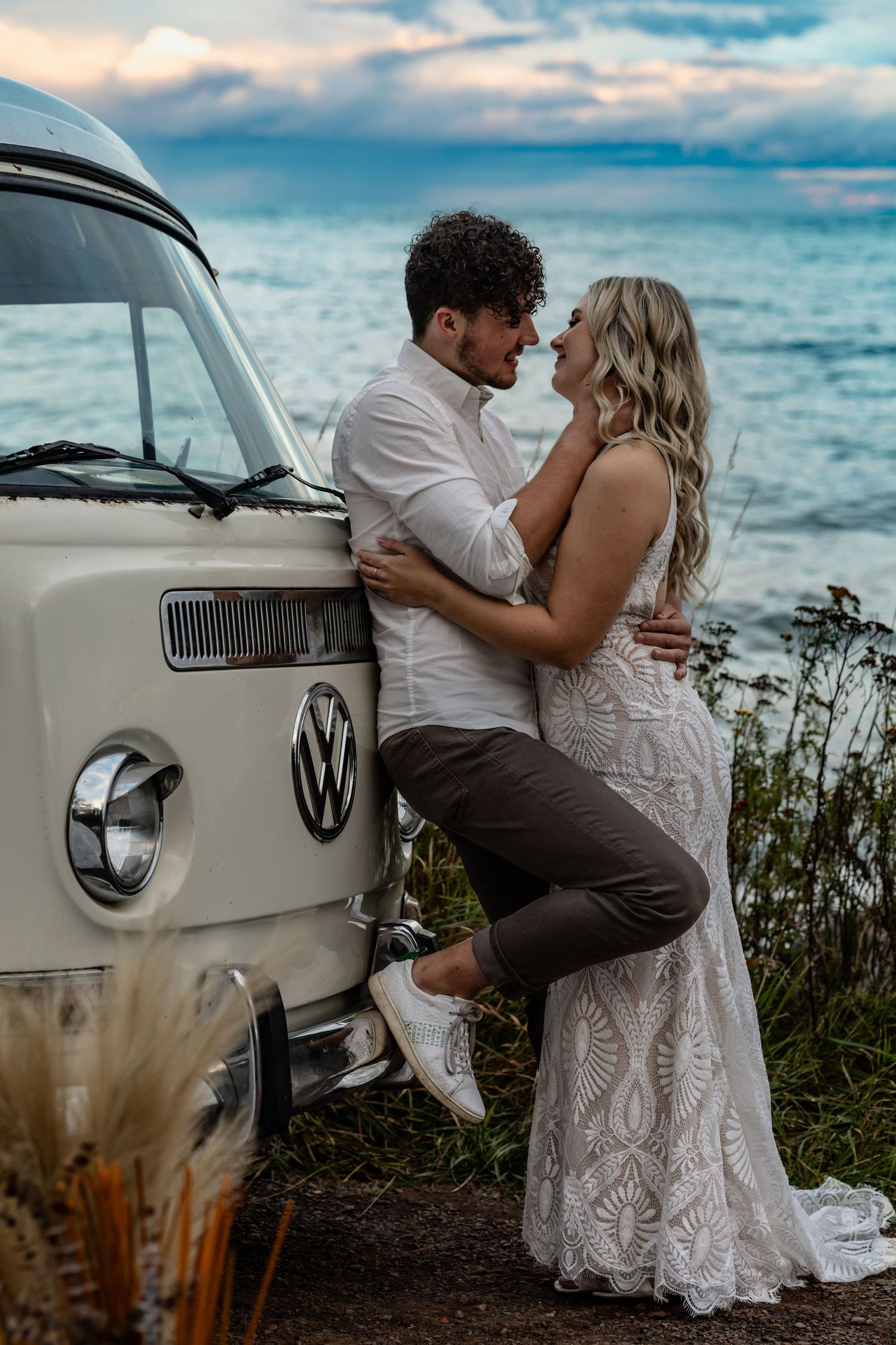 Couple sharing a romantic moment leaning against a vintage VW van by the ocean at sunset, bride in lace gown.