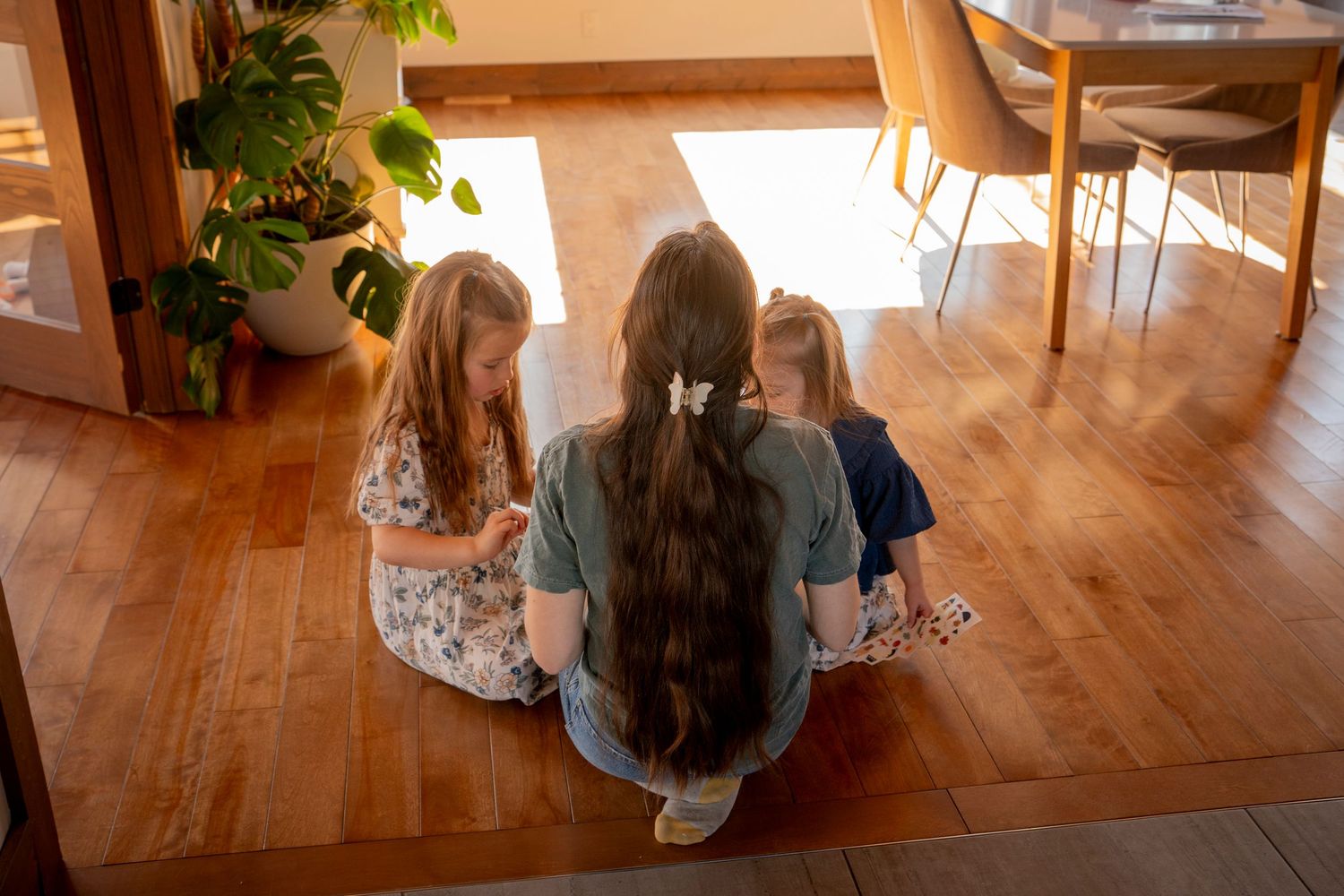 Teen girl and two young sisters sit on hardwood floor playing with cards together in bright home.