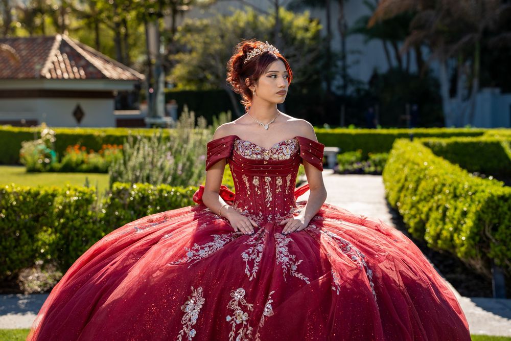 Young woman in elegant red off-shoulder quinceañera ballgown with white lace embroidery posing in a lush garden.
