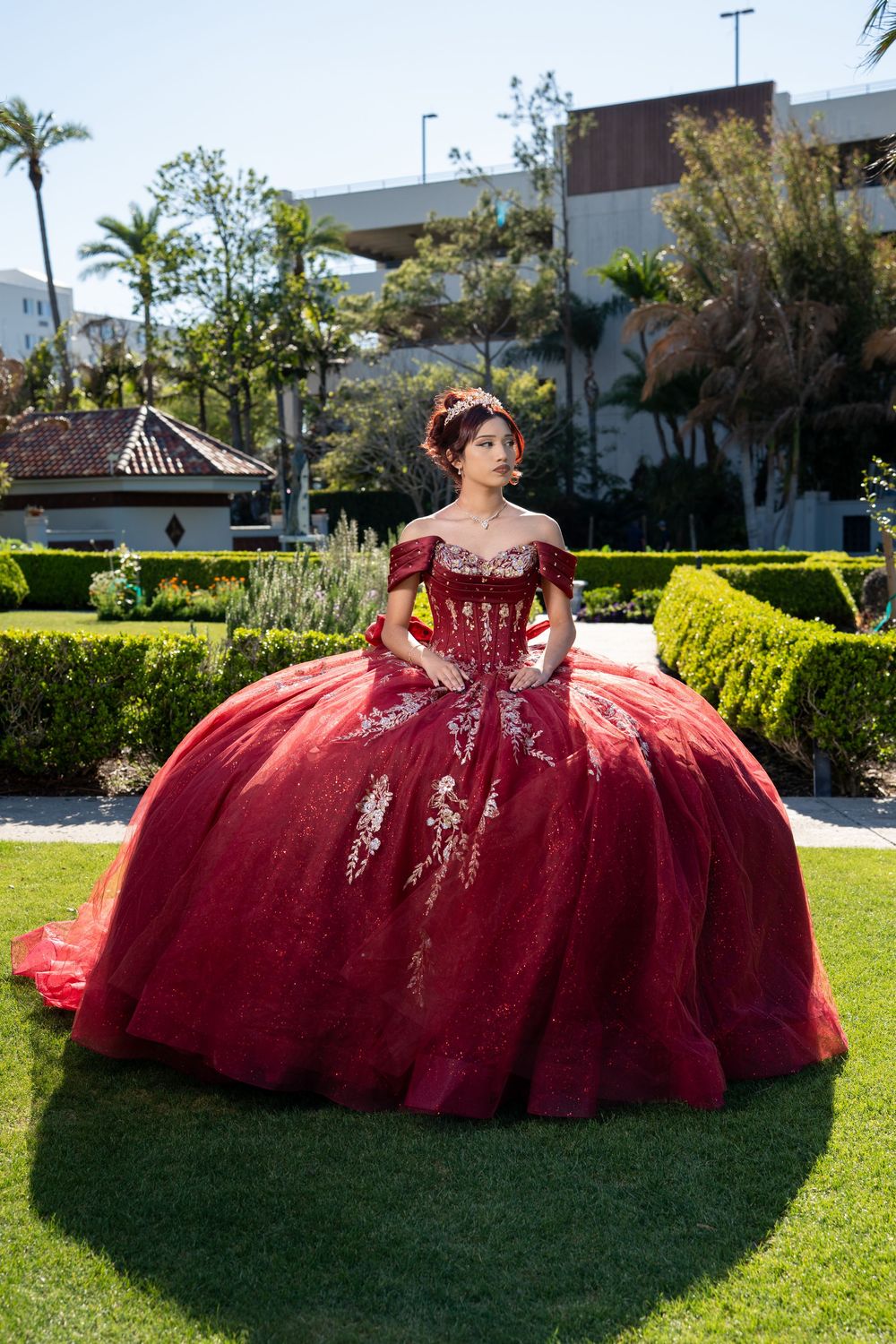 Young woman in a stunning red ball gown with gold embroidery poses in a lush garden for her quinceañera.
