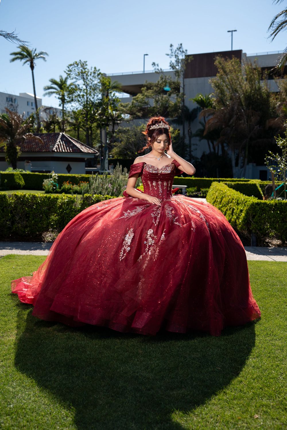 Young woman in a stunning red quinceañera ball gown with floral embroidery poses in a lush garden setting.