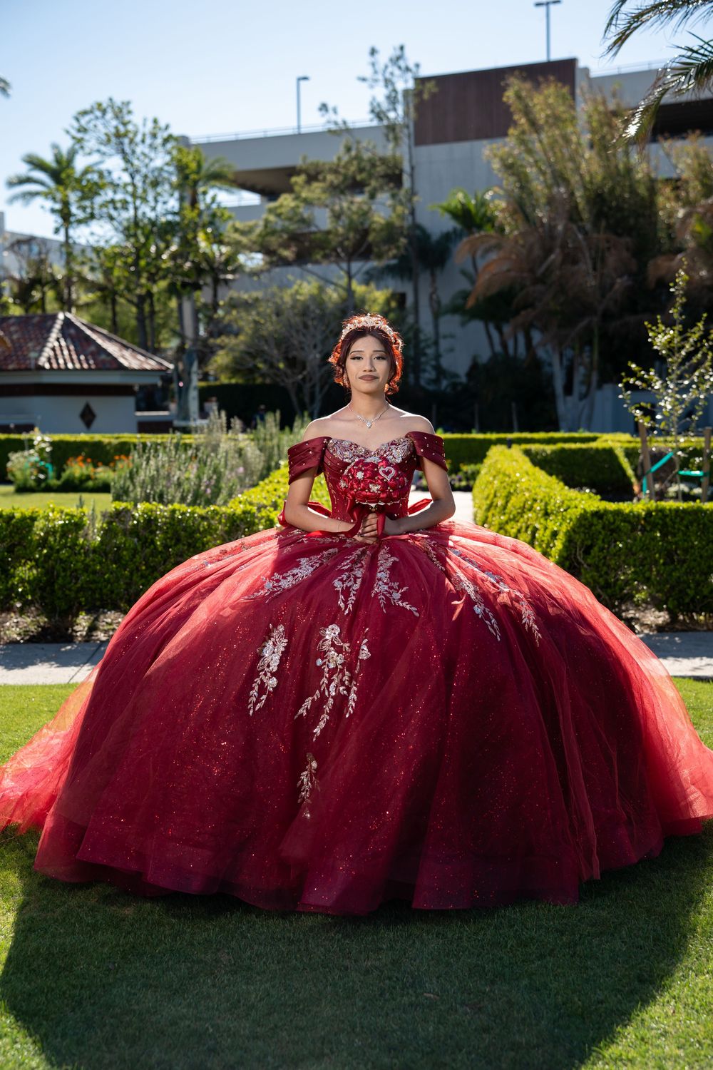 Young woman in a stunning red quinceañera ball gown with silver embroidery poses in a lush garden setting.