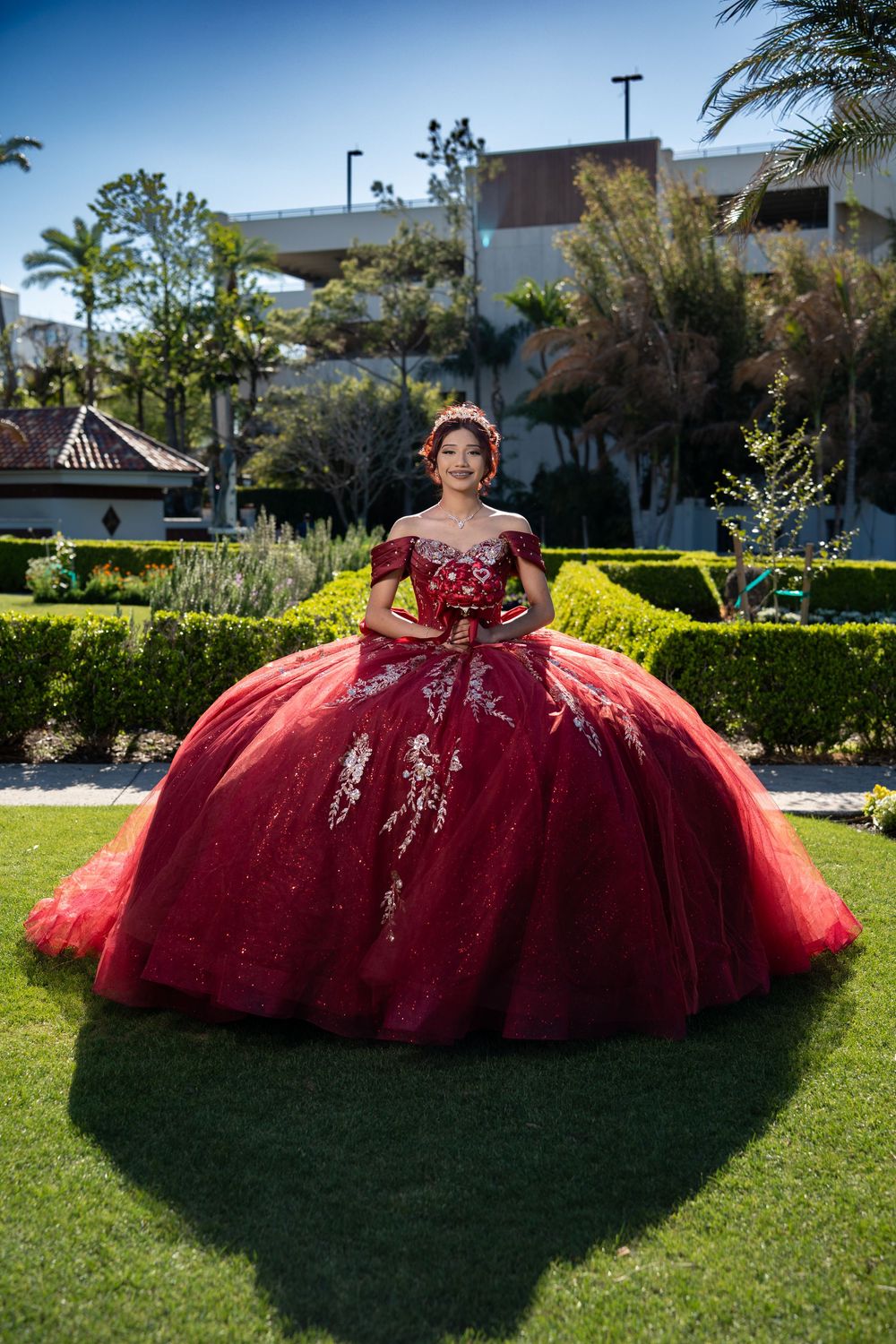 Young woman in a stunning red ballgown with gold embroidery poses in a lush garden for her Quinceañera.