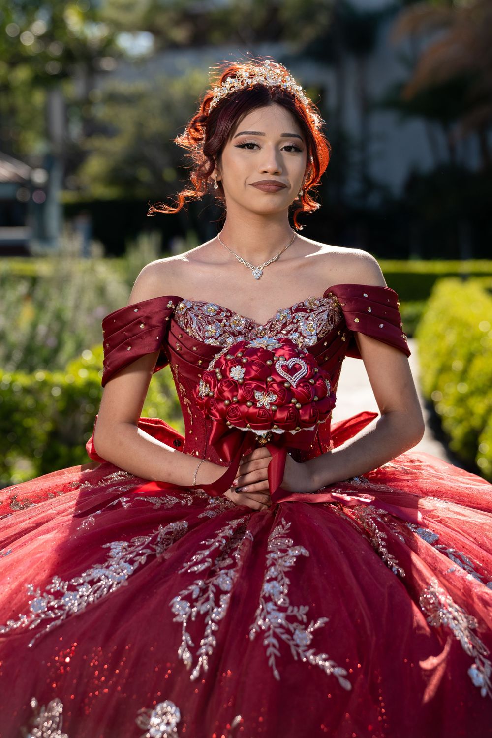 Young woman in red quinceañera gown with floral embroidery, crown, and heart brooch posing outdoors.
