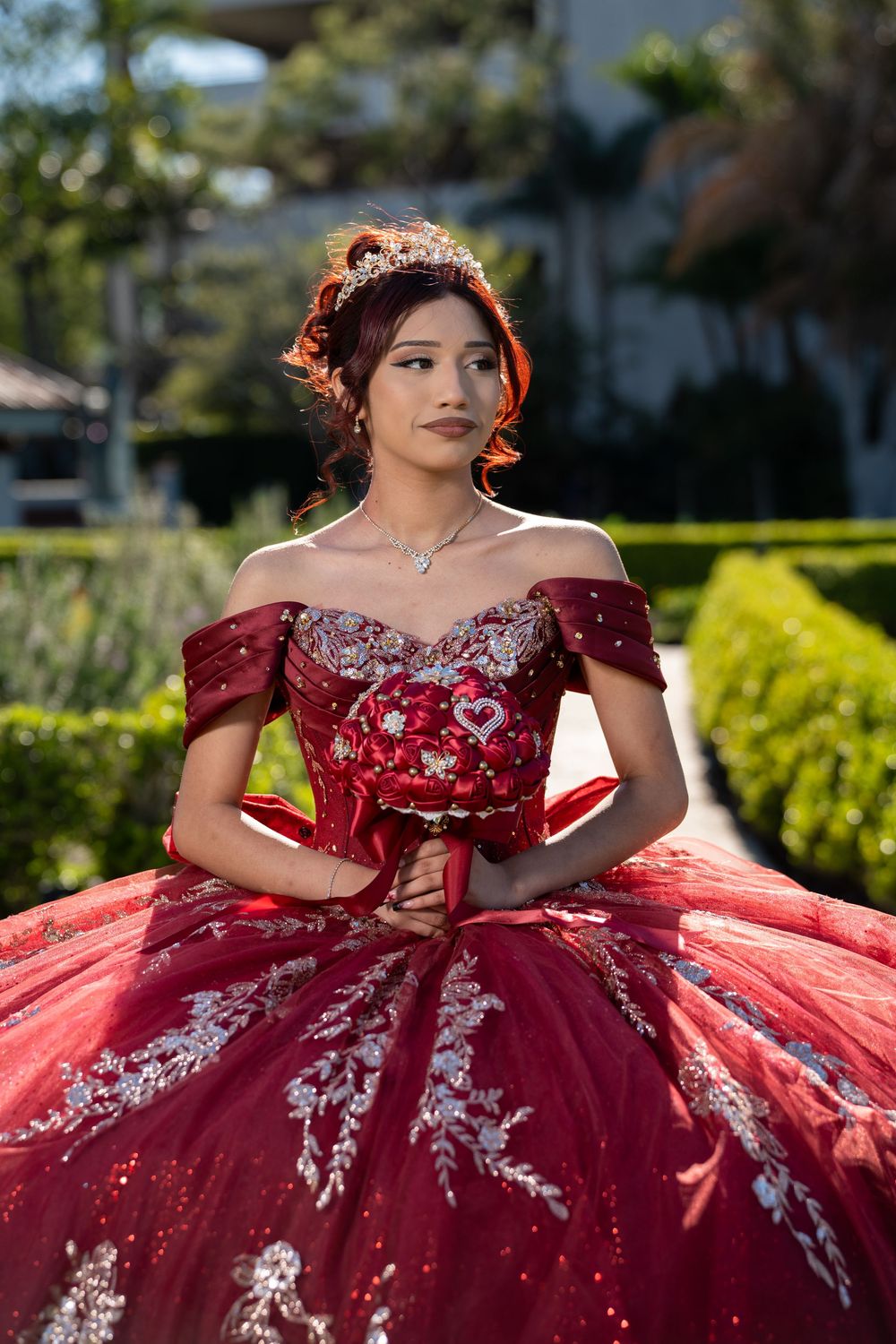 Young woman in a red off-shoulder quinceañera ball gown with silver embroidery and tiara poses outdoors.