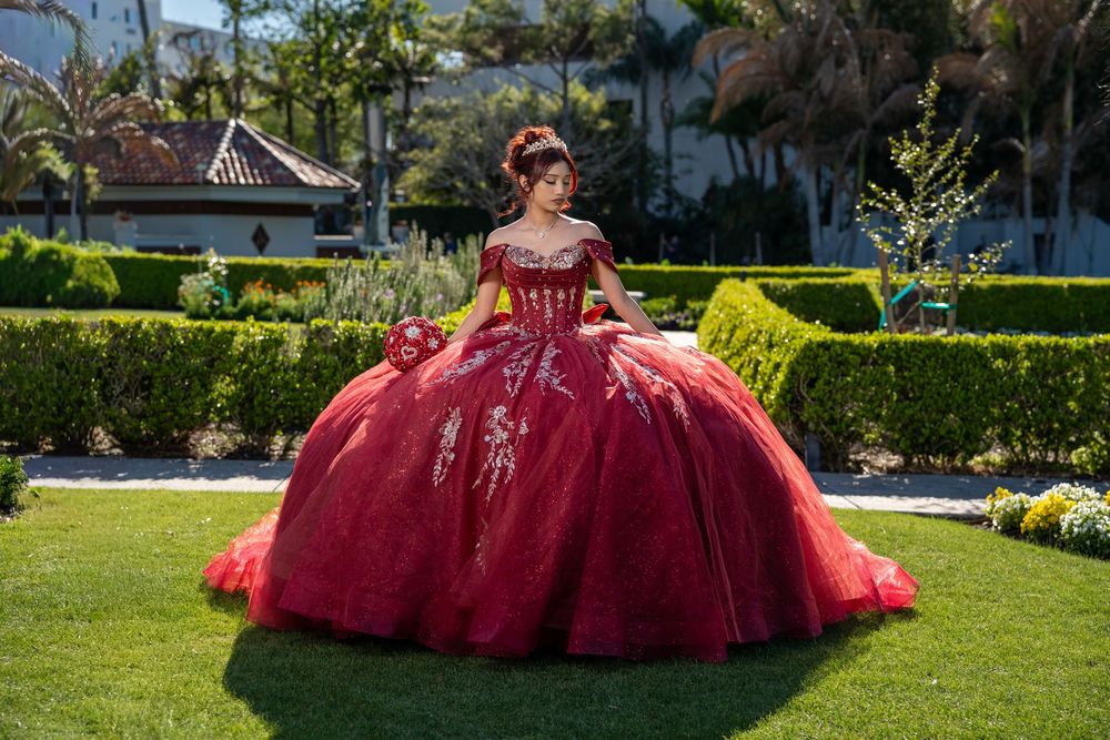 Young woman in a stunning red ball gown quinceañera dress posing in a lush garden setting.