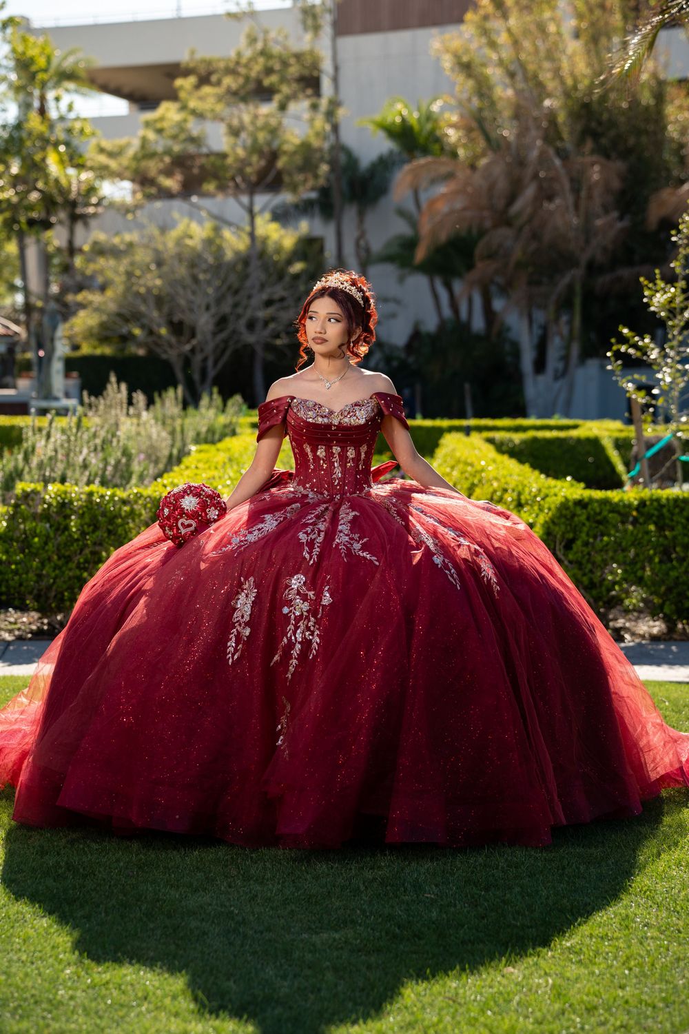 Young woman in a stunning red ball gown with silver embroidery poses in a garden for her Quinceañera.
