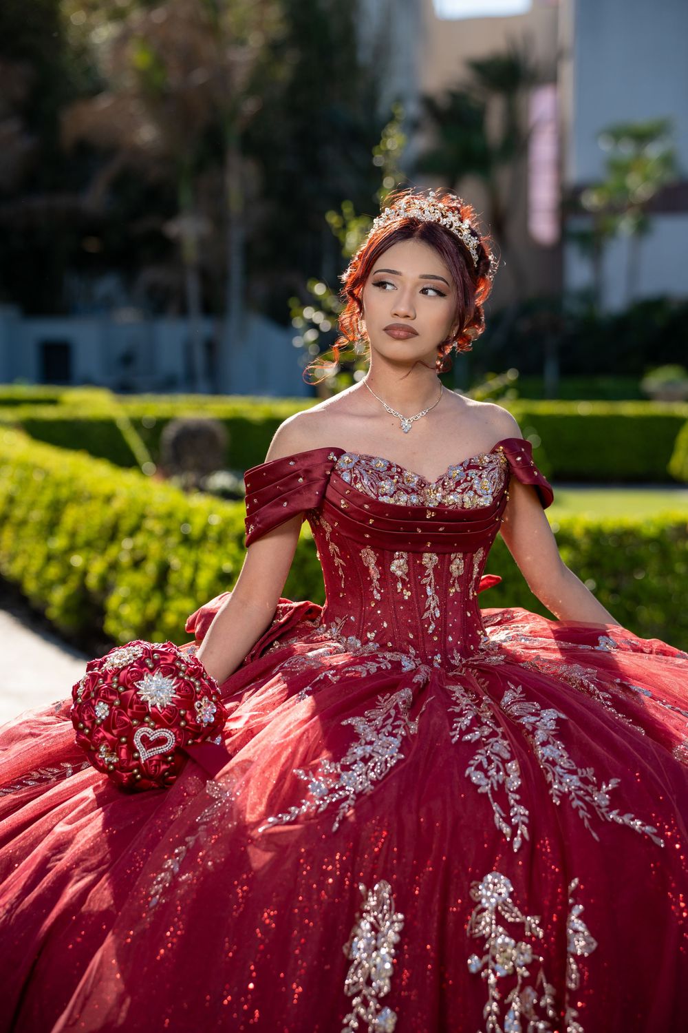 Young woman in red quinceañera ball gown with silver embroidery, floral crown, and bouquet posing in a garden.