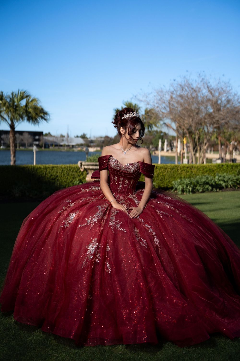Young woman in a stunning red ball gown with gold embroidery and tiara poses outdoors near a waterfront.