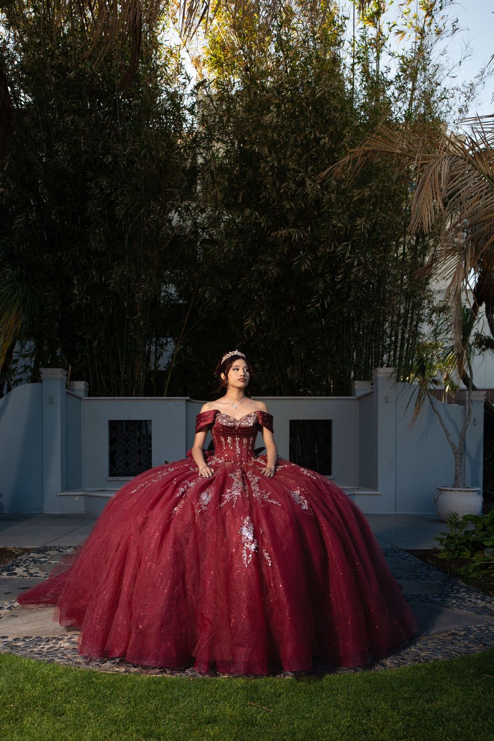 Young woman in a stunning burgundy ball gown with silver embroidery poses in an elegant garden setting.