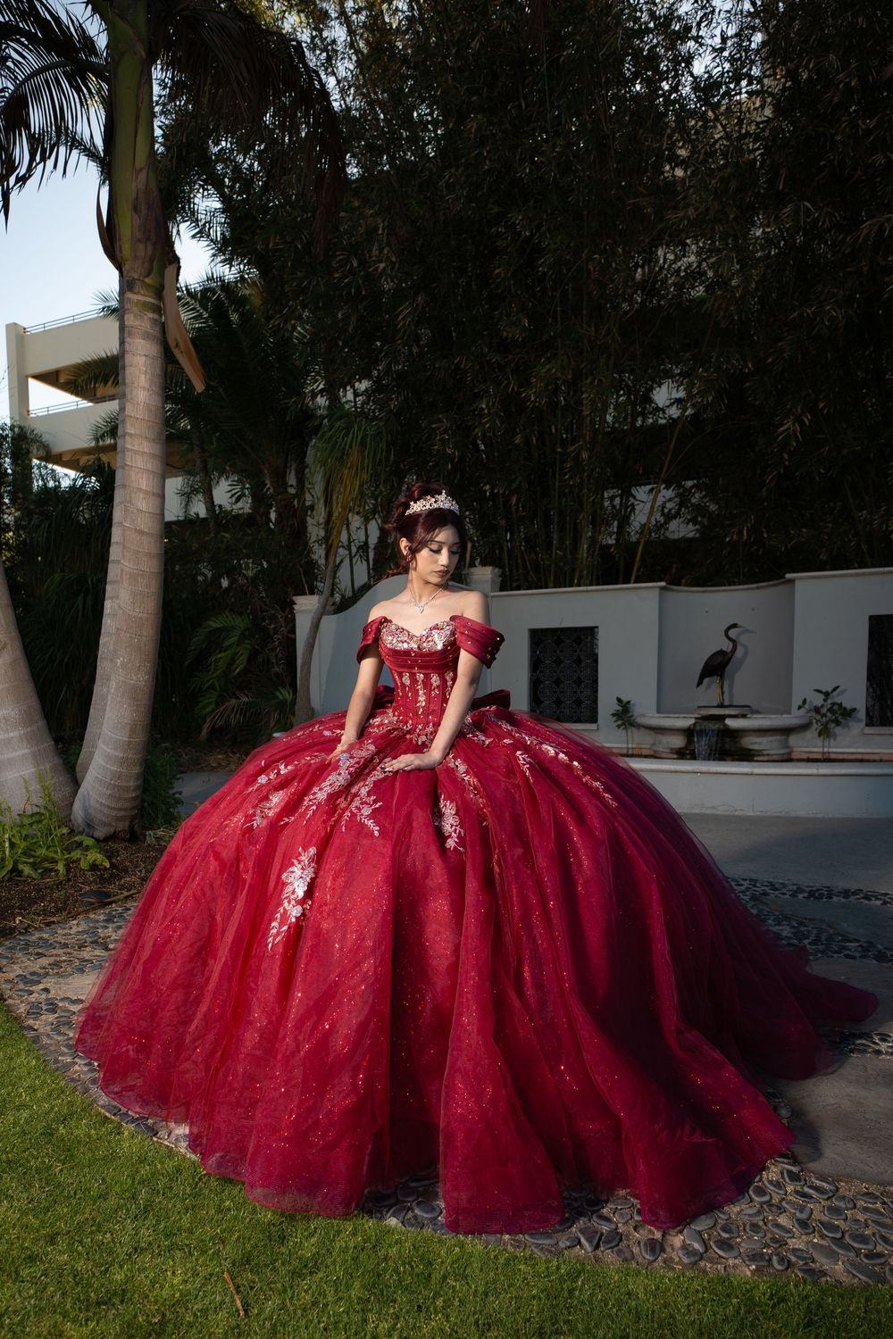Young woman in a stunning red ball gown quinceañera dress with floral embroidery, posing in a lush garden setting.