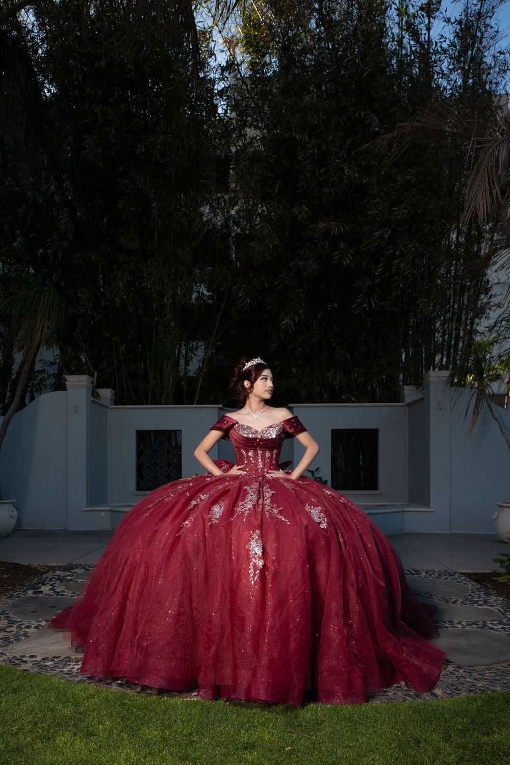 Young woman in a stunning crimson ball gown with gold embroidery posing outdoors for quinceañera photos.