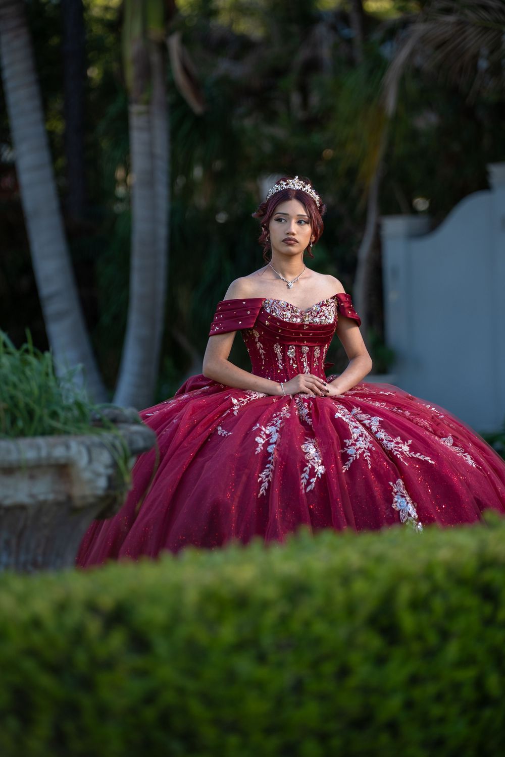 Young woman in a stunning crimson ball gown with silver embroidery and tiara poses outdoors for quinceañera photos.