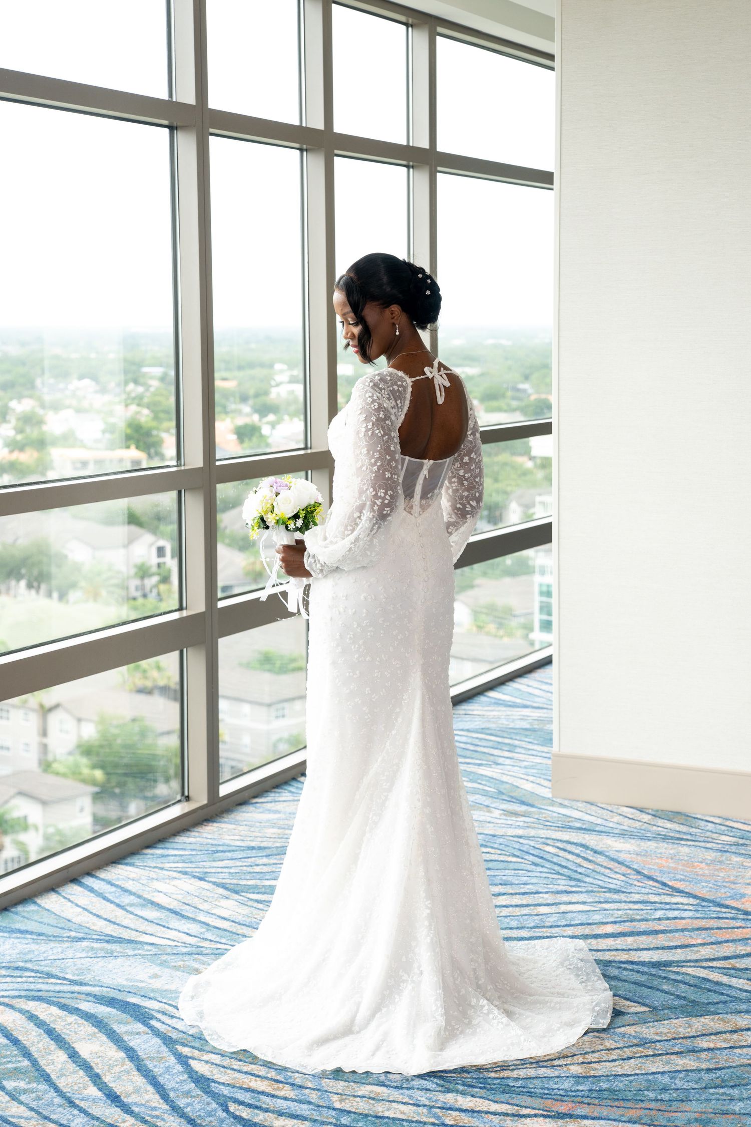 Elegant bride in a long-sleeve lace wedding gown holding a bouquet, standing by large windows with a city view.
