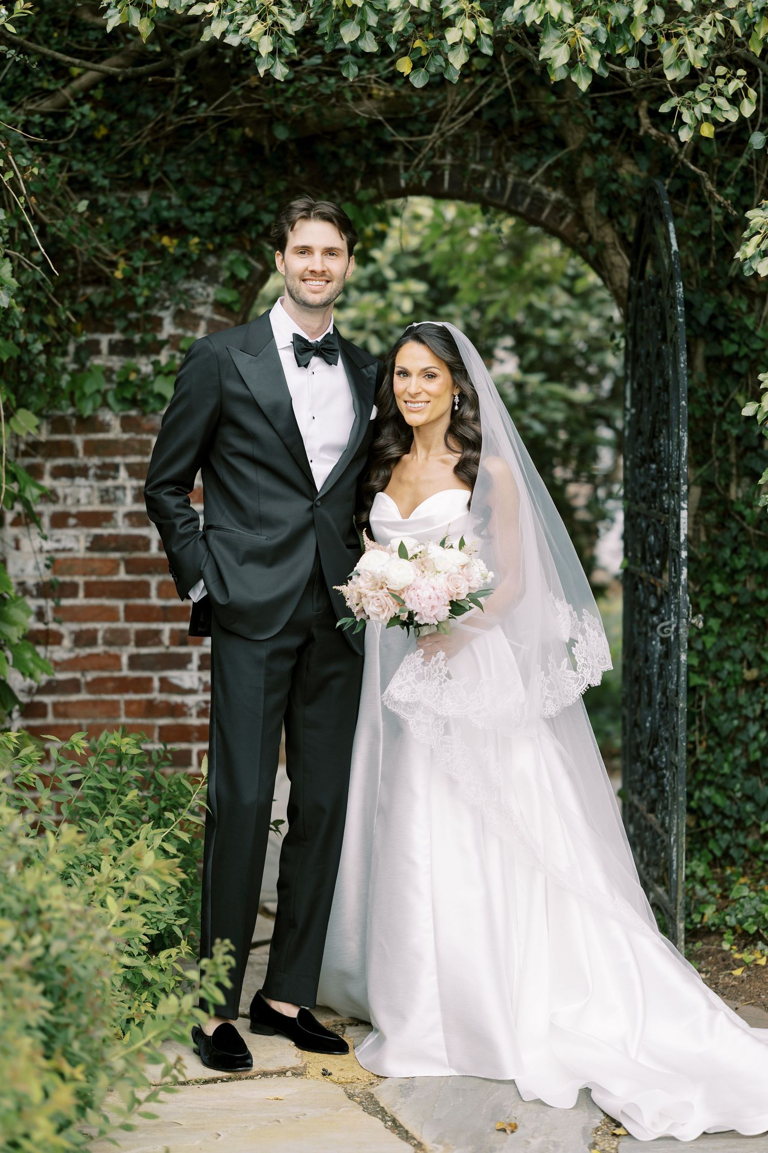 Bride and groom posing under ivy-covered arch, she in white ballgown with veil, he in black tuxedo with bow tie.