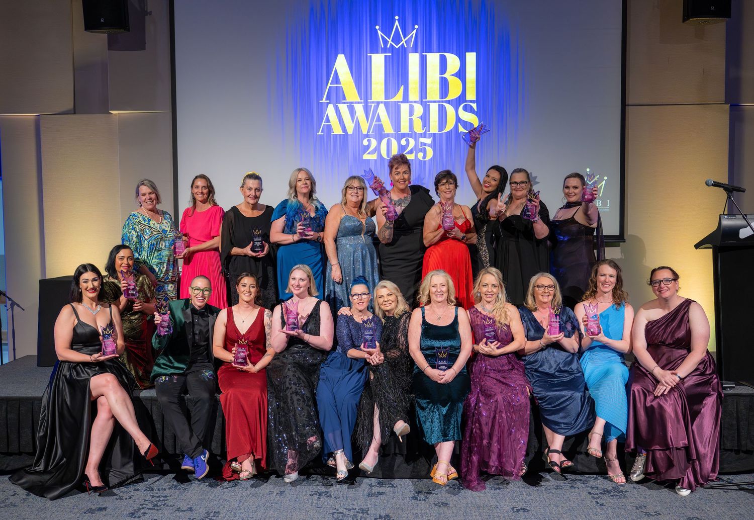 Group of women posing together at the Alibi Awards 2025 ceremony, holding trophies on a decorated stage.
