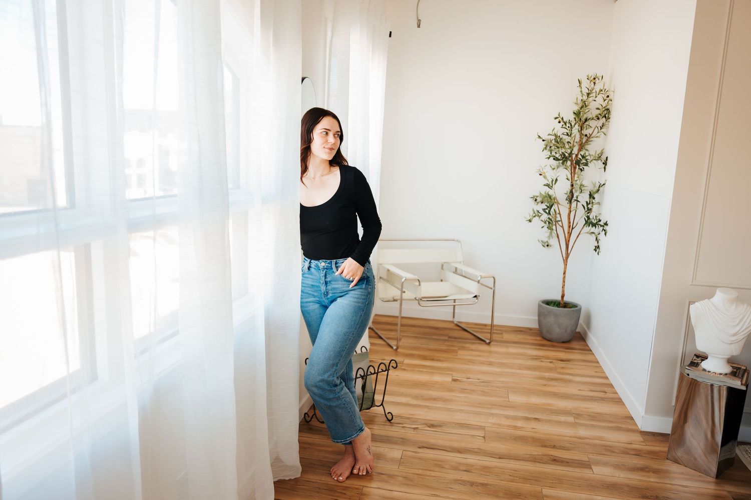 Woman in a tshirt and jeans leans against a bright white wall in a modern studio in St.Albert. Lifestyle photography branding poses.