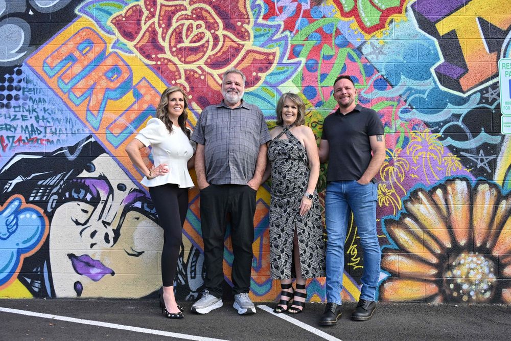 Four people smiling in front of a vibrant colorful graffiti mural wall with flowers and abstract art.