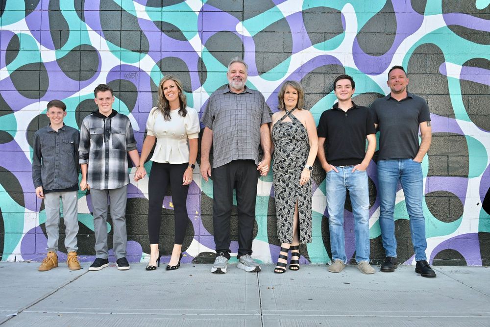 Seven people posing together in front of a colorful teal and purple abstract mural wall outdoors.
