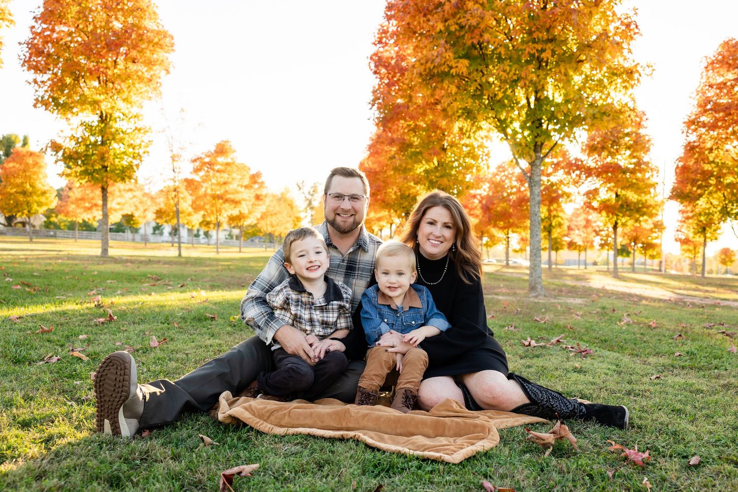 A group sits together on a blanket in a park during autumn with vibrant orange trees in the background.