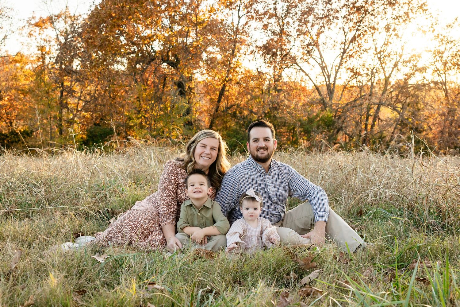 A family sits together in a golden autumn field at sunset with tall grass and trees in warm fall colors.