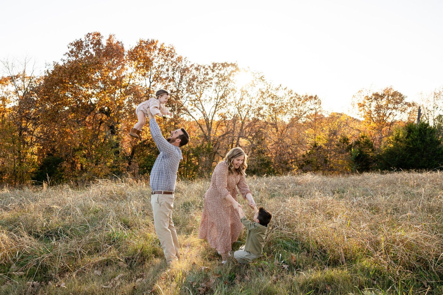 A couple walks through a golden field at sunset holding hands while wearing casual autumn attire.