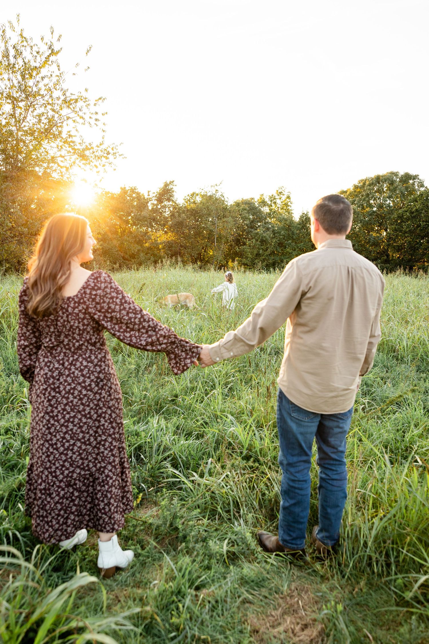 A couple walks hand in hand through a sunlit meadow at golden hour, surrounded by tall grass and wildflowers.