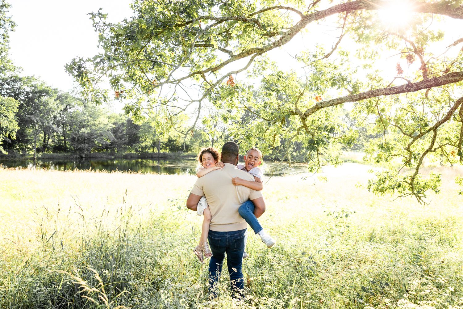 Two people embrace in a sunlit meadow surrounded by tall grass and trees during golden hour.
