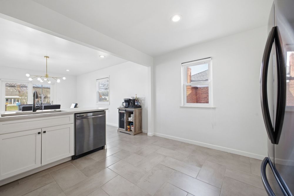 Modern white kitchen with stainless dishwasher, tile floors, chandelier, and bright windows.
