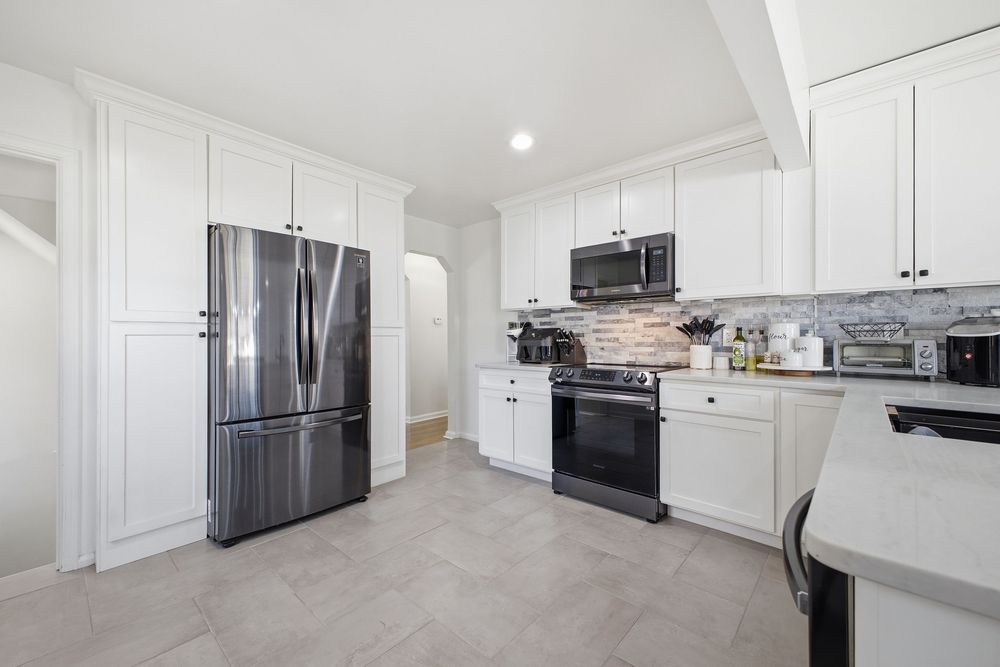 White kitchen featuring stainless steel refrigerator, gas range, microwave, and stone tile backsplash.