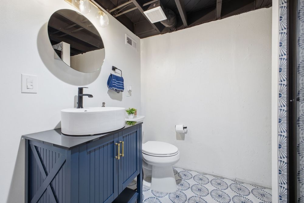 Basement bathroom with navy blue vanity, vessel sink, round mirror, and patterned tile floor.