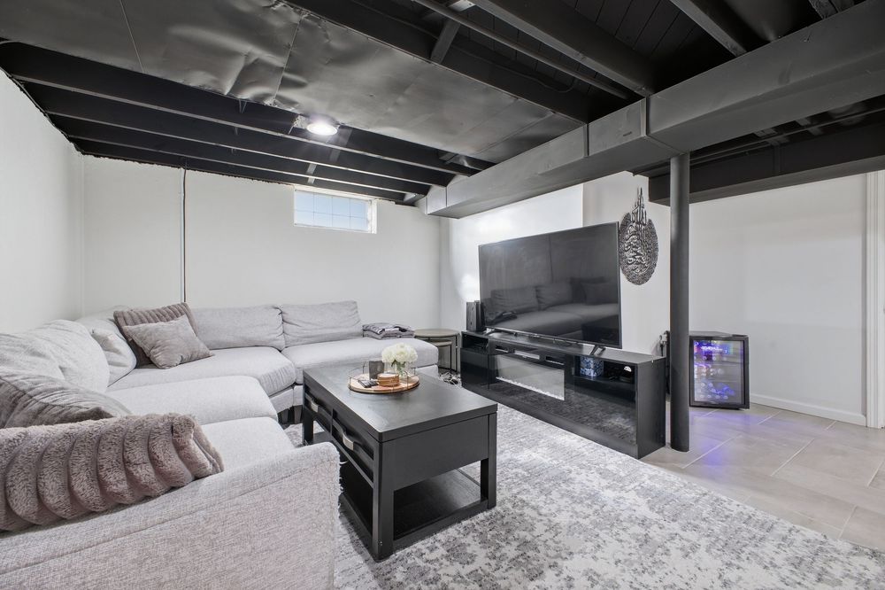 Modern basement living room with gray sectional sofa, black coffee table, wall-mounted TV, and exposed black ceiling beams.