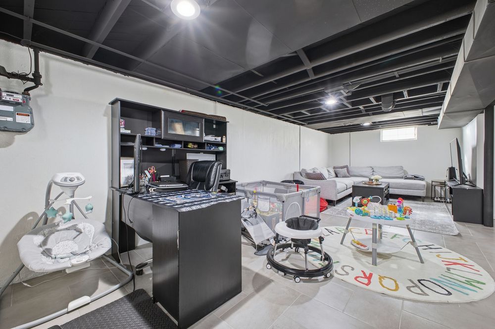 Spacious basement multipurpose room with black desk, shelving, play area rug, and exposed black ceiling joists.