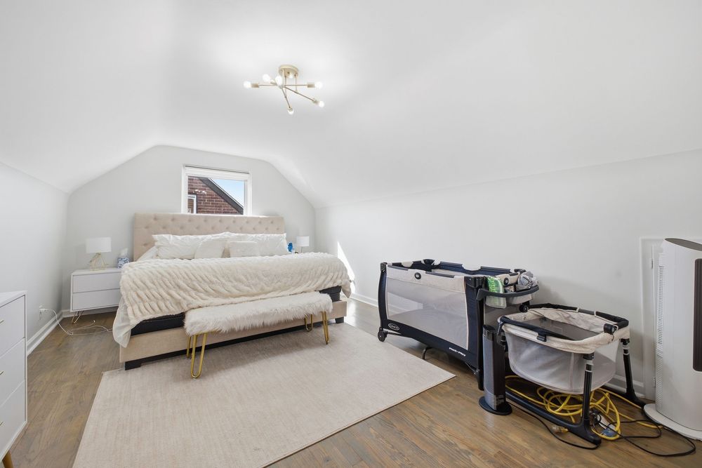 Bright attic bedroom with vaulted white ceiling, beige upholstered bed, hardwood floors, and portable crib.