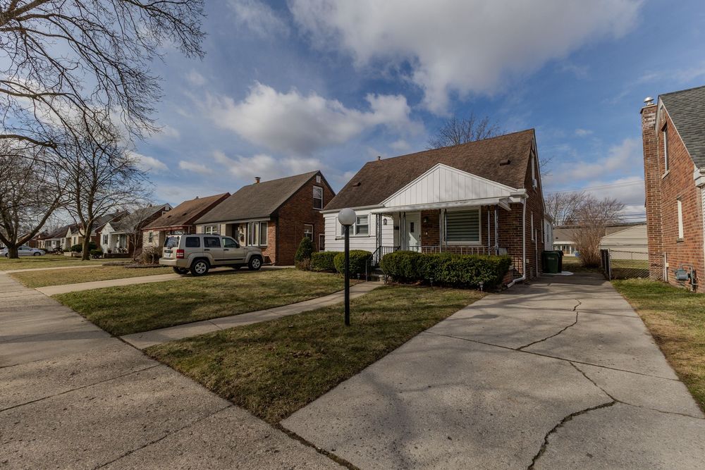 Suburban bungalow home with white siding, brown roof, concrete driveway, and neighboring brick houses on tree-lined street.