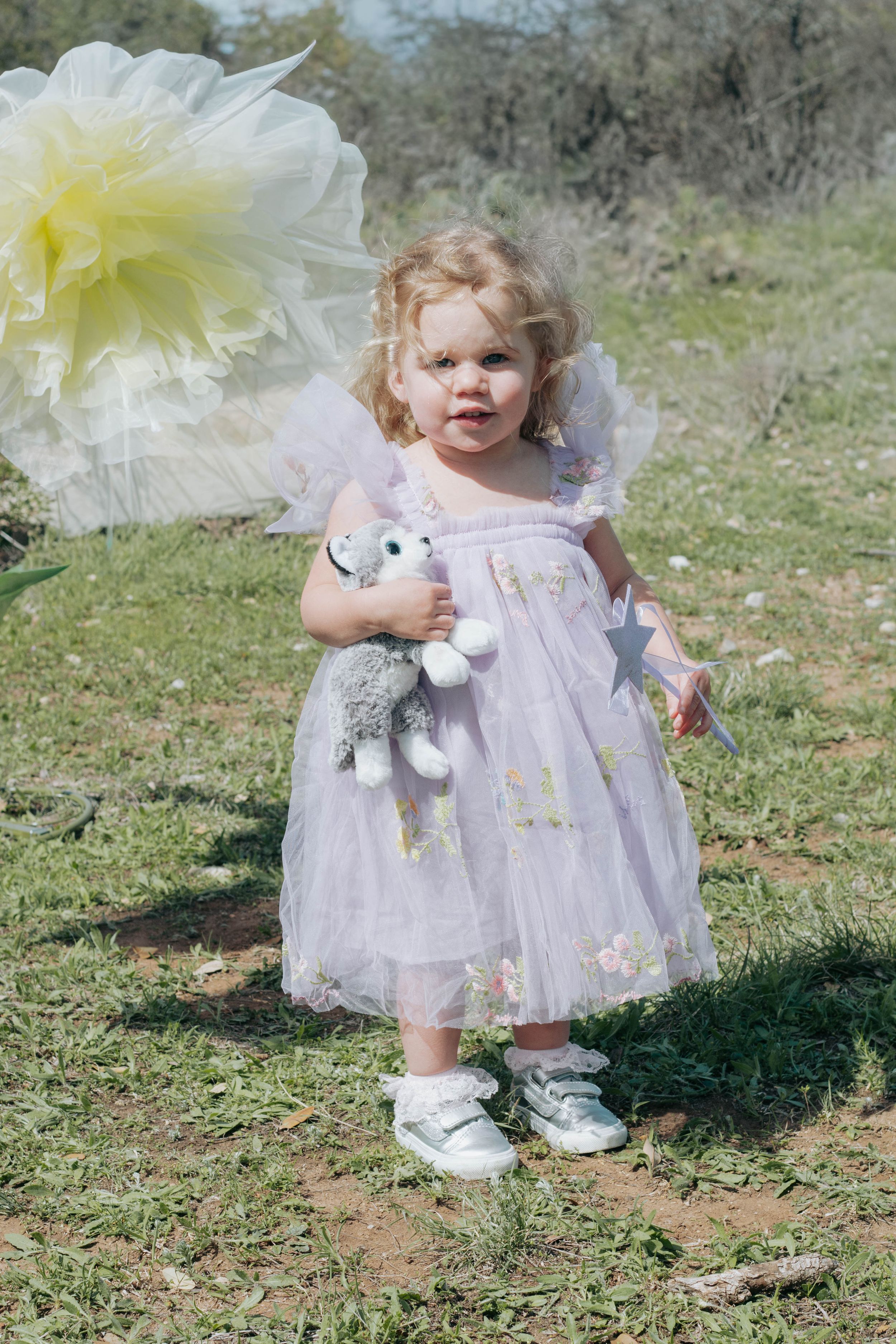 Toddler girl in lavender tulle fairy dress holding stuffed animal toy, standing outdoors on grass in white sneakers.